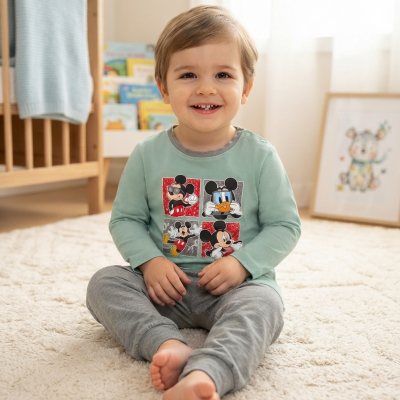 Bebé sentado no chão com camisola verde água com estampado Mickey e calças cinzentas.