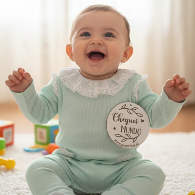 Bebé com fato macaco verde água e gola branca sentada em tapete branco com placa Cheguei MUNDO