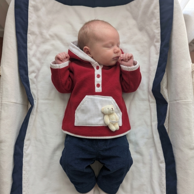 Bebé dormindo vestido com camisa vermelha com bolso e ursinho e calças azuis, deitado em lençol branco com bordas azuis