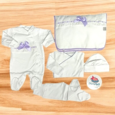 Conjunto de roupa de bebé branca com laço lilás e manta, em superfície de madeira