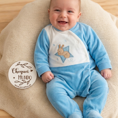 Bebé sorridente vestido com macacão azul e branco com coelho, deitado em manta bege e placa de madeira com texto