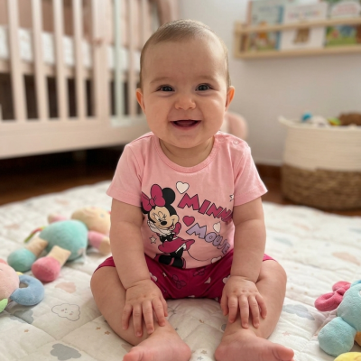Bebé sorridente com roupa rosa de Minnie Mouse sentado em tapete almofadado com berço branco e brinquedos ao fundo.