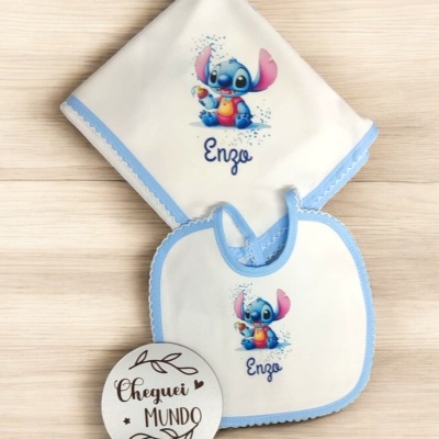 Babete e fralda de bebé com desenho de personagem azul e rosa e texto 'Enzo', sobre mesa clara com placa 'Cheguei MUNDO'.
