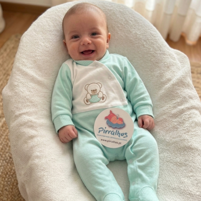 Bebé a sorrir de macacão verde claro com ursinho bordado, deitado em almofada branca