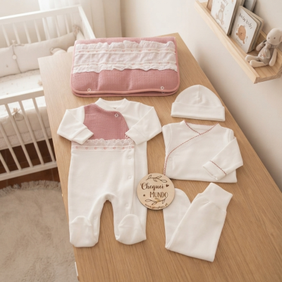 Conjunto de roupa de bebé creme e rosa com renda e manta acolchoada numa mesa de madeira ao lado de um berço e estante infantil