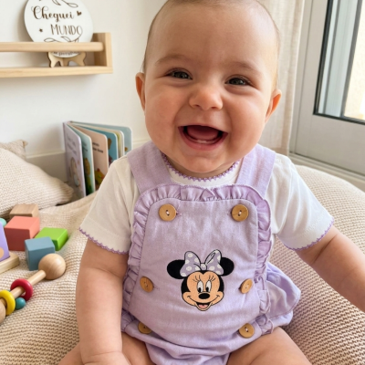 Bebé sorridente com babygrow lilás da Minnie e t-shirt branca num quarto com brinquedos.