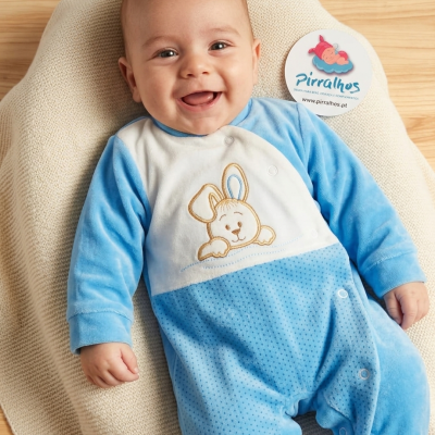 Macacão de bebé azul e branco com bordado de coelho, bebé sorridente deitado numa almofada bege