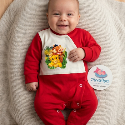 Bebé a sorrir vestido com pijama vermelho com painel branco estampado com personagens do Rei Leão, deitado num almofadão bege