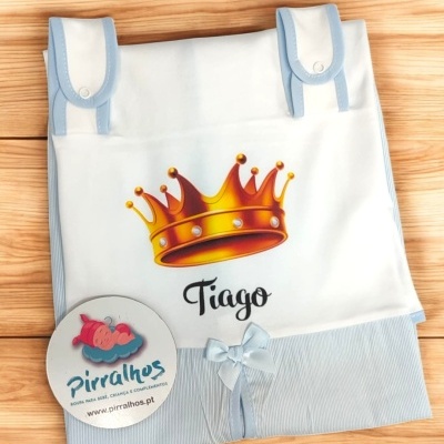 Capa para bebé branca com coroa amarela e o nome Tiago, detalhes em azul claro, sobre superfície de madeira
