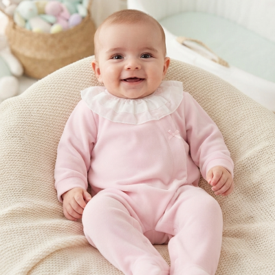 Bebé sorridente vestido com pijama rosa, sentado numa almofada bege de malha em quarto claro com cesta de brinquedos ao fundo