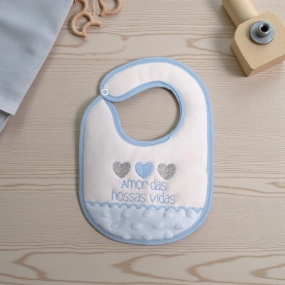 Babete de bebé branco com bordas azuis e texto bordado colocado sobre mesa de madeira clara.