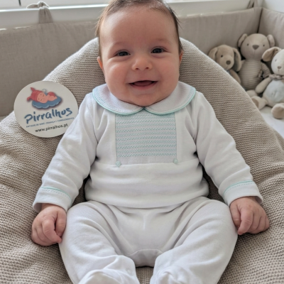 Bebé vestido com macacão branco de gola arredondada com detalhes azuis sentado em almofada bege com peluches ao fundo