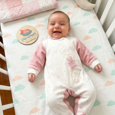 Bebé sorridente de macacão rosa e branco a deitar numa cama com lençol de nuvens e cobertor rosa e bege