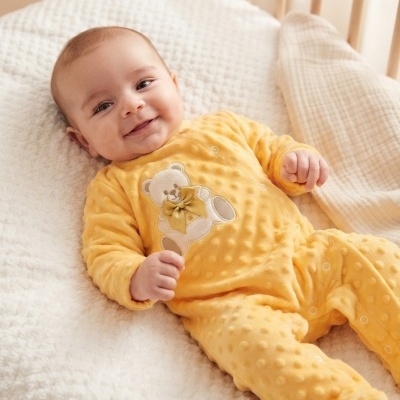 Bebé sorridente deitado em cama com fato amarelo texturizado com ursinho bordado.