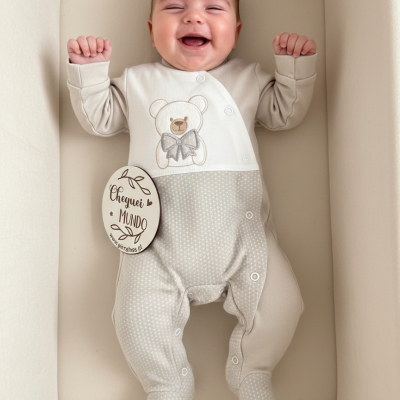 Bebé sorridente de macacão cinzento com bolinhas brancas e ursinho bordado, deitado numa almofada creme.