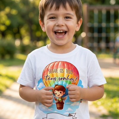 Camisola branca infantil com estampa de balão de ar quente e texto em português numa imagem ao ar livre.