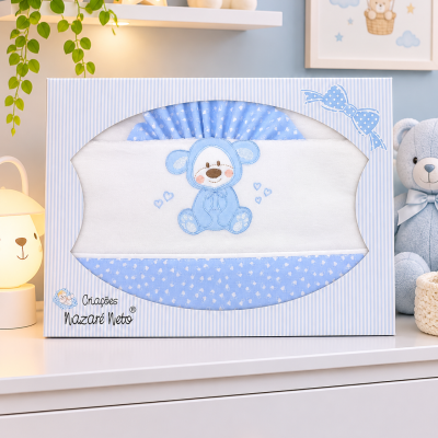 Conjunto de roupa de bebé azul e branca com ursinho bordado numa caixa decorada, sobre uma cómoda branca.