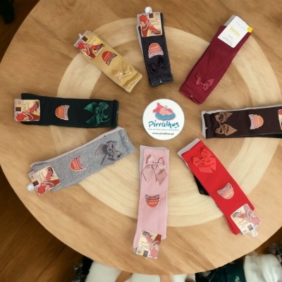 Oito pares de meias coloridas com laços dispostas em círculo sobre mesa de madeira e etiqueta redonda no centro.