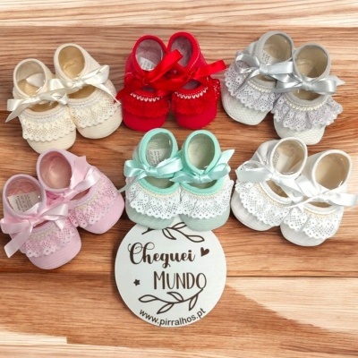 Seis pares de sapatinhos de bebé coloridos com laços e renda, dispostos em círculo com cartão central sobre madeira clara.