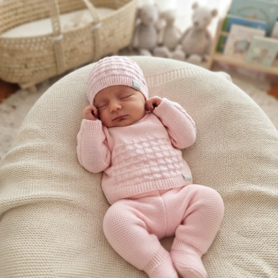 Bebé dormindo com roupa de malha rosa clara e gorro, em almofada bege