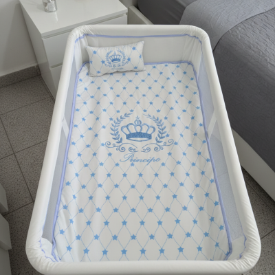 Berço branco com colchão e almofada decorados em azul, ao lado de mesa de cabeceira branca com difusor e candeeiro