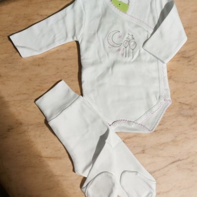 Conjunto de roupa de bebé branco com bordado e calças de pé sobre superfície marmoreada