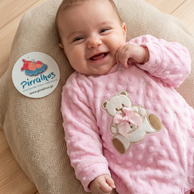 Bebé a sorrir com macacão rosa texturado e urso branco com laço cor-de-rosa deitado em almofada bege sobre chão de madeira