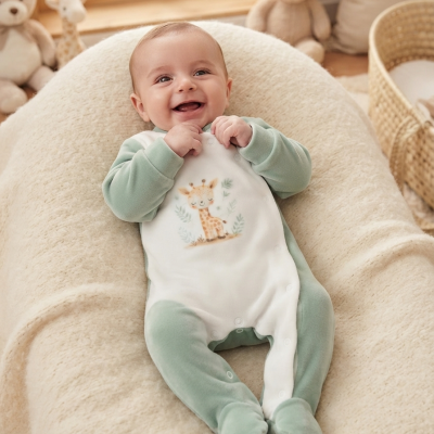 Bebé sorridente deitado com macacão verde e branco com estampa de girafa numa almofada cremosa, rodeado de peluches e cesto de ráfia.