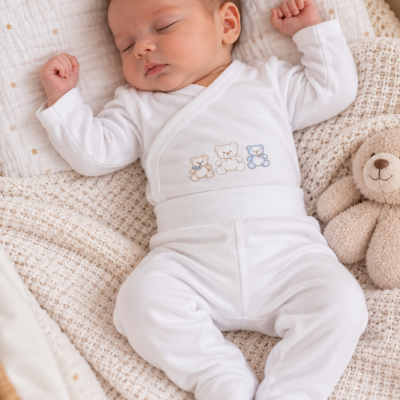 Bebé a dormir com pijama branco e ursinhos bordados, sobre mantas claras, com peluche de urso ao lado.