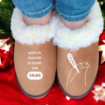 Chinelos castanhos forrados com pelúcia branca e texto impresso, usados por uma pessoa com calças de ganga, sobre superfície vermelha com decoração.