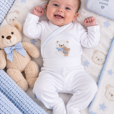Bebé numa manta com ursinhos e estrela, vestido com macacão branco bordado, ao lado de ursinho de peluche.