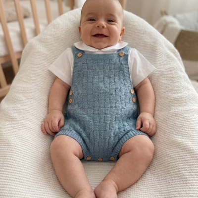 Bebé sorridente com babygrow azul claro e t-shirt branca sentado numa almofada cremosa.