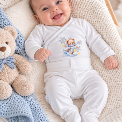 Bebé com macacão branco com estampa de urso a fazer snowboard, deitado no berço com manta bege e urso de peluche com laço azul