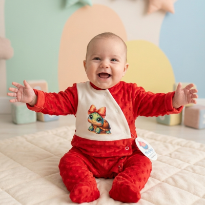 Bebé sorridente com fato vermelho e babete branco com estampa de tartaruga, sentado em tapete creme com fundo colorido e brinquedos.