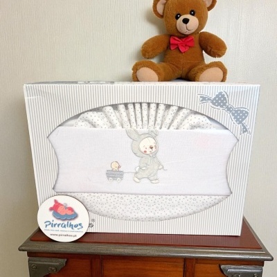 Conjunto roupa bebé branco com coelhinho bordado, em caixa decorada, sobre móvel de madeira com ursinho de peluche castanho.