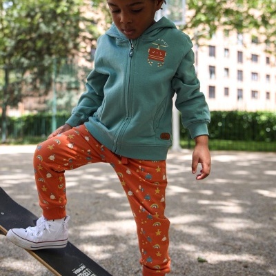 Criança com casaco azul, calças laranjas estampadas e ténis brancos num skate