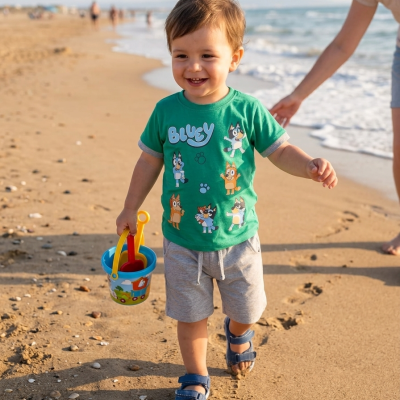 Criança com camisola verde BLUEY, calções cinza e sandálias azuis na praia a segurar balde de plástico azul