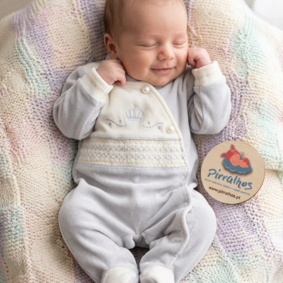 Bebé vestido com macacão cinzento claro com detalhes creme e coroa bordada, a dormir na manta de malha multicolorida com etiqueta da Pirralhos