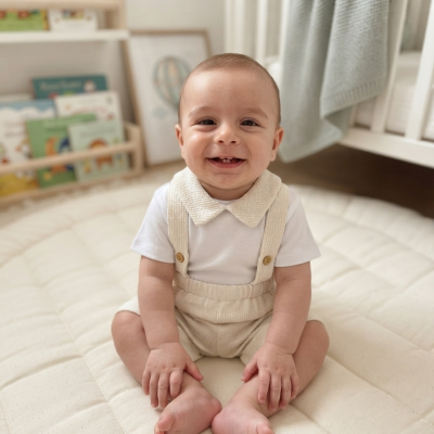 Bebé sentado com roupa branca e bege num quarto com estante de livros e berço.