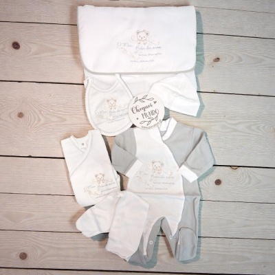 Conjunto de roupa de bebé com bordado de urso e frase, em tons de branco e cinzento, sobre fundo de madeira clara.