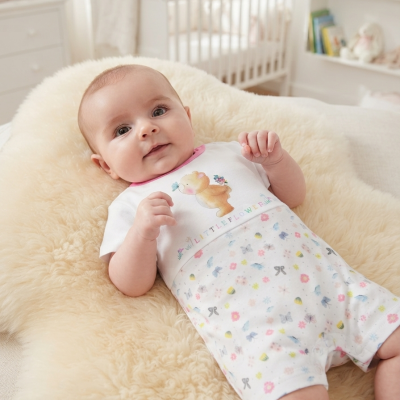 Babygrow branco com padrão floral e imagem de urso, bebé deitado em almofada creme num quarto de bebé.