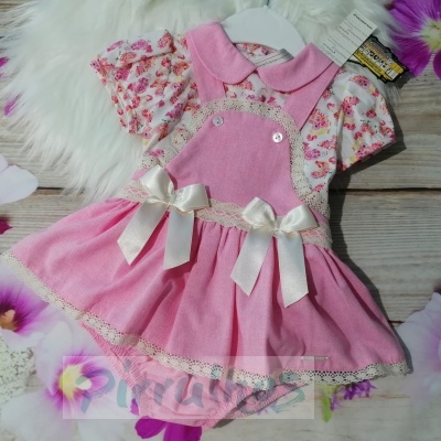 Vestido rosa para bebé com padrão floral, laços e detalhes em renda creme numa superfície de madeira com flores artificiais.