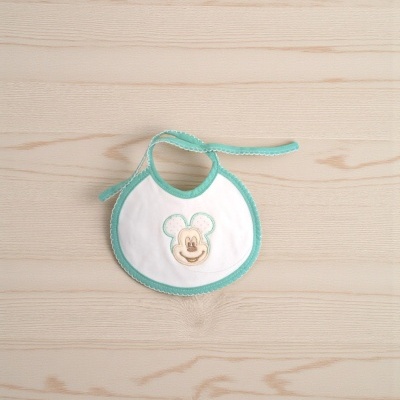 Babete branco e verde água com rosto bordado de rato sobre superfície de madeira clara