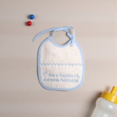 Babete branco com bordado azul e utensílios de bebé em fundo bege