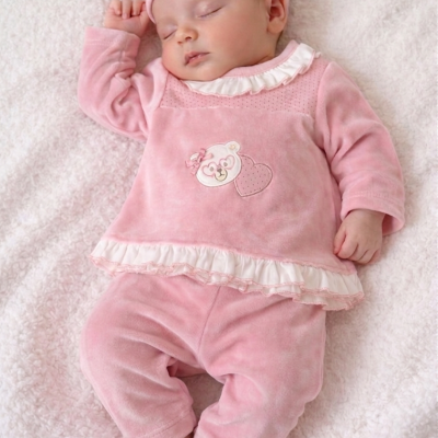 Bebé recém-nascida vestindo roupa rosa felpuda com ursinho e coração, deitada em cobertor branco felpudo