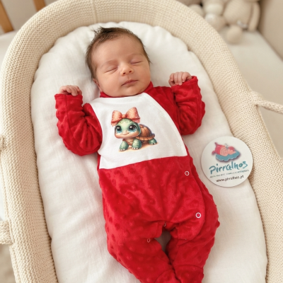 Bebé vestido com um macacão vermelho e branco com uma tartaruga estampada, deitado numa almofada branca dentro de uma cesta bege.