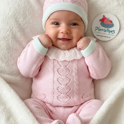 Bebé com conjunto em malha rosa com gola branca e gorro, sobre manta branca, com selo de marca Pirralhos.