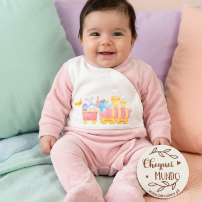 Bebé sorridente com pijama rosa e branco com estampa de comboio, sentado em almofadas coloridas segurando placa com texto.