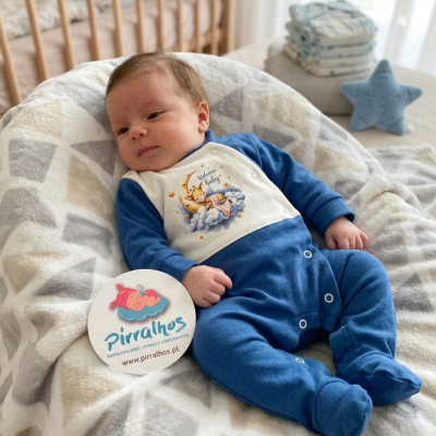 Bebé vestido de azul e branco com estampa, deitado num berço acolchoado cinza e bege com etiqueta da marca Pirralhos