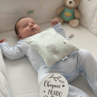 Bebé de pijama riscado azul e branco no berço com almofada estampada e ursinho peluche.